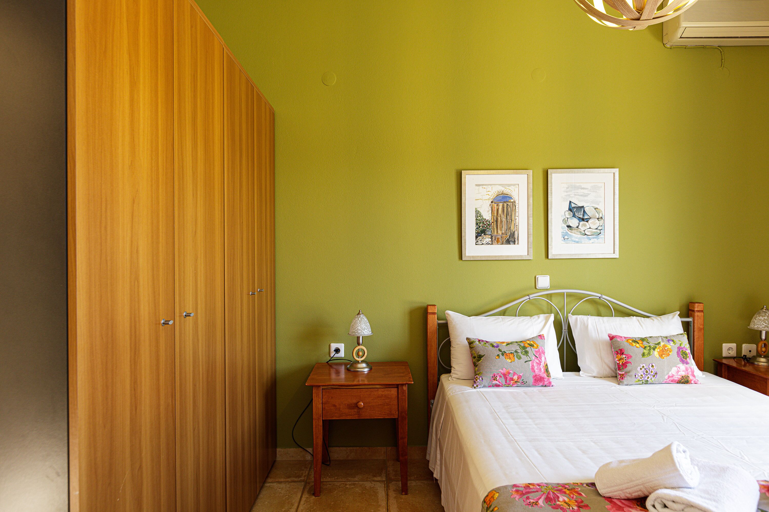 A bedroom with a wooden wardrobe, a bed with floral bedding, and two framed artworks on the wall.