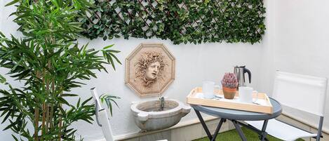 A small outdoor seating area with a table, chairs, a potted plant, a stone fountain, and a decorative wall.