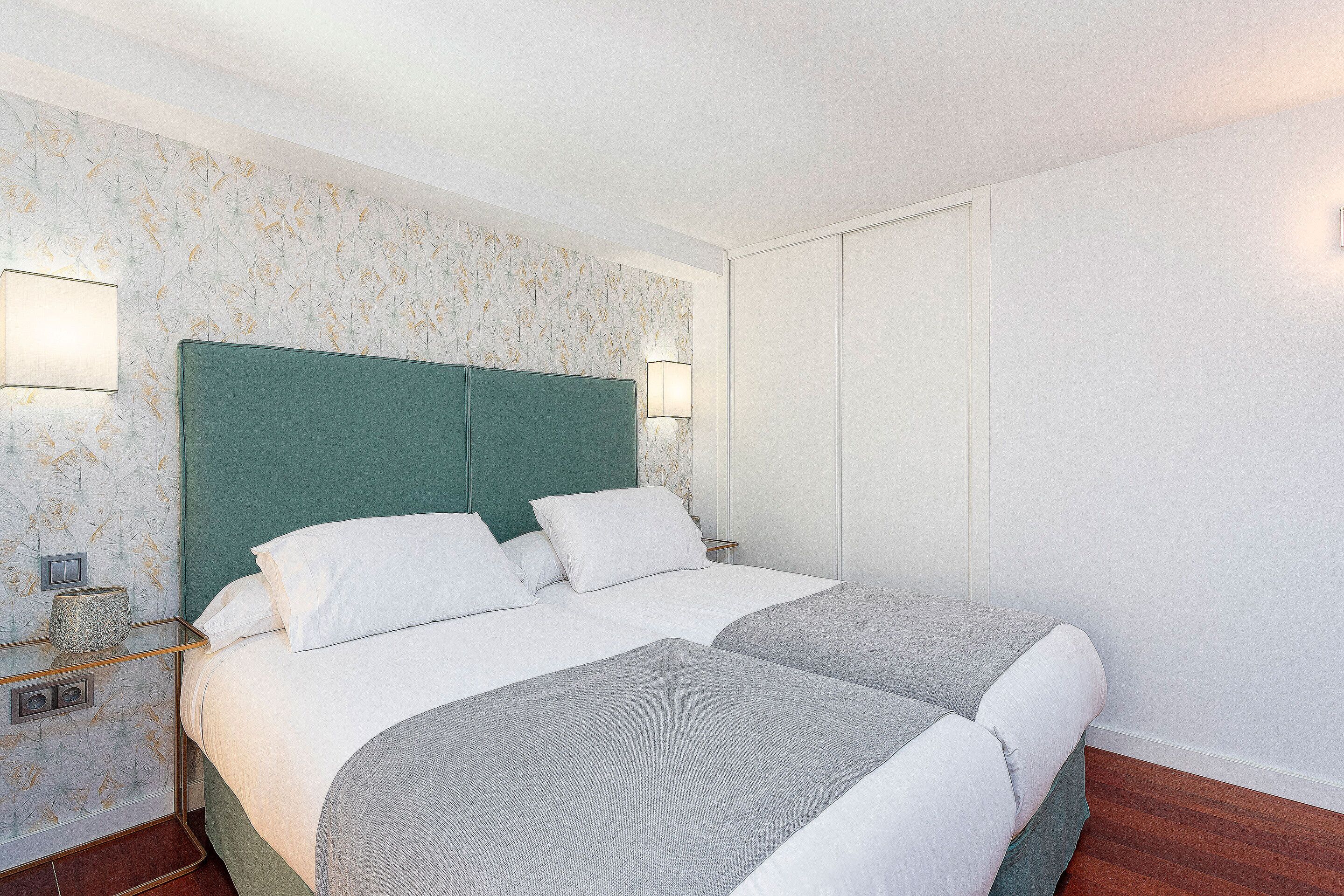 A double bed with a green headboard, white bedding, and a grey throw blanket. There are two bedside tables with lamps. The room has a floral wallpaper and a wooden floor.