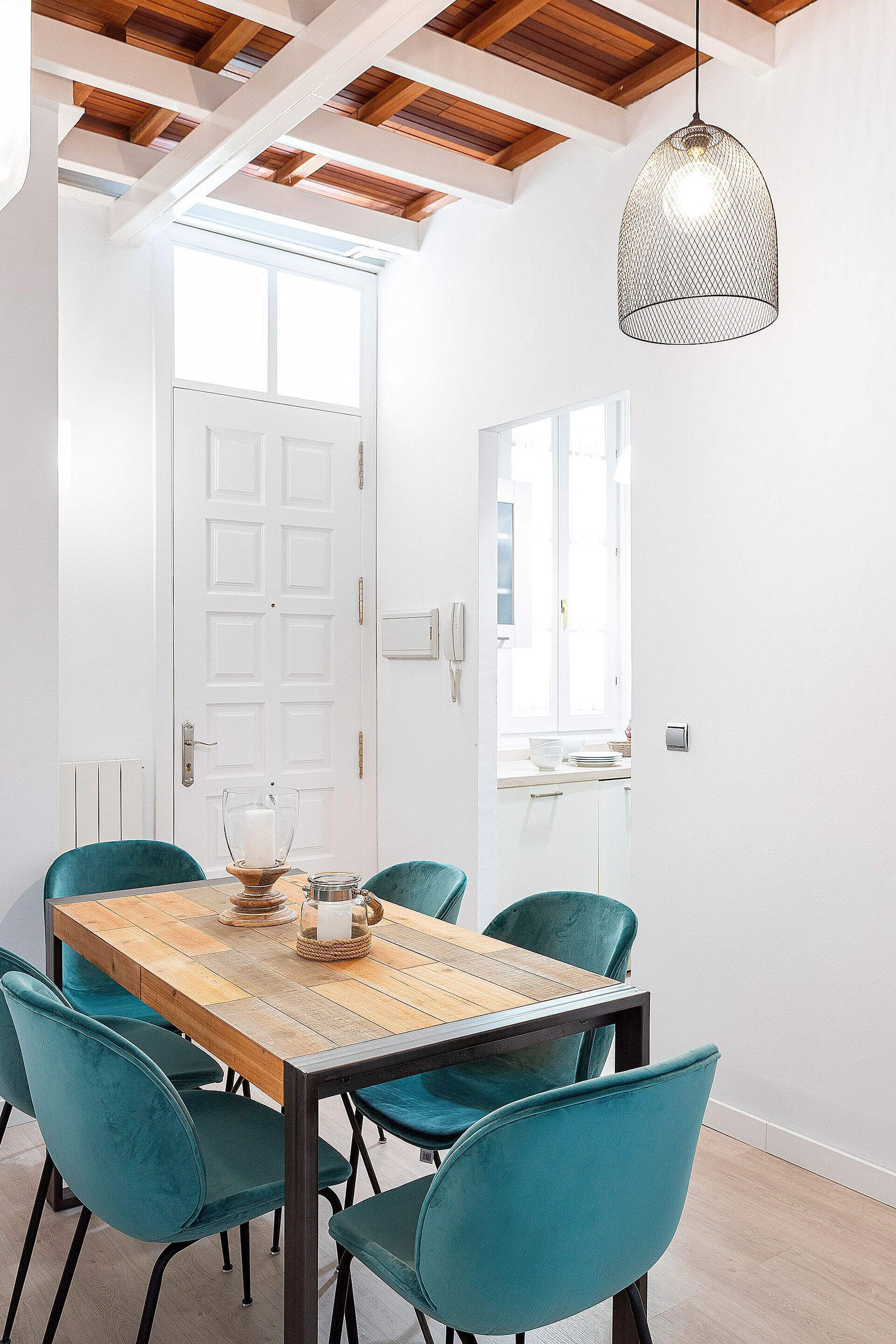 Un comedor con mesa de madera y sillas azul verdosas, una lámpara colgante y una puerta blanca con ventanal.