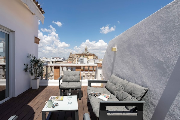 A rooftop terrace with a white wall, a sofa, a chair, a table, and a potted plant.