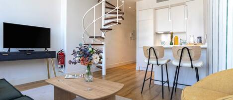 A modern living room with a flat-screen TV, a wooden coffee table, and a spiral staircase leading to a kitchen area.