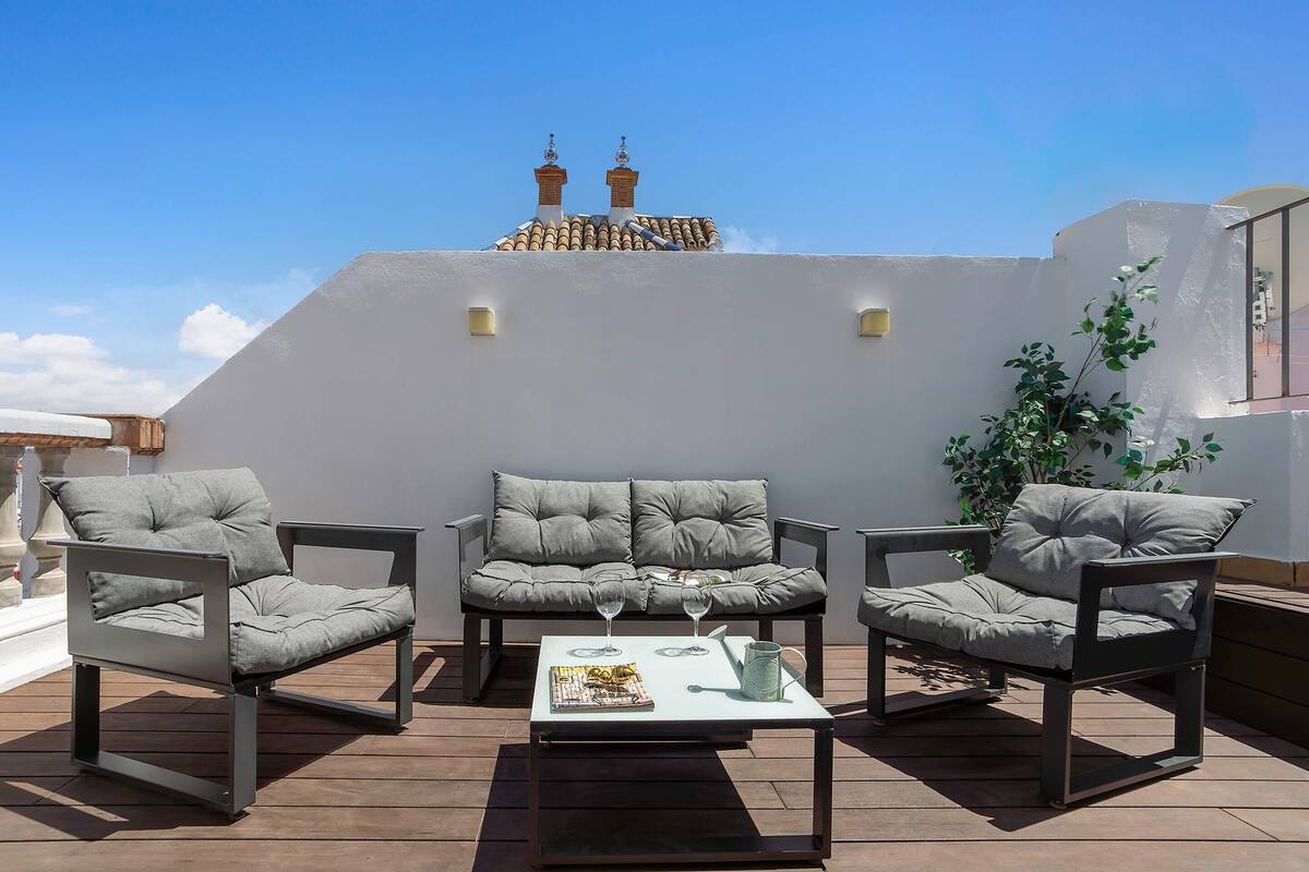 A rooftop patio with a sofa set, a table with glasses and a pitcher, and a view of a building with chimneys.