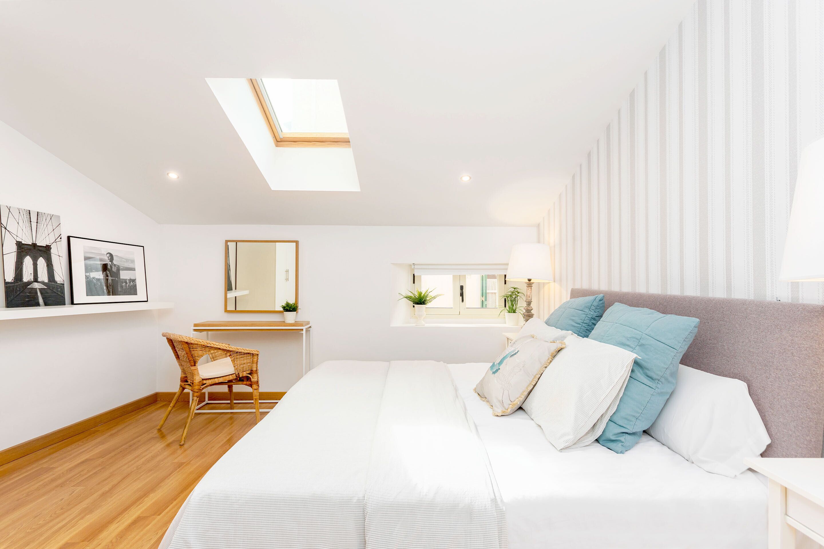 Quarto moderno com claraboia, cama com roupa de cama branca, cadeira de vime, mesa de cabeceira, espelho e janela com plantas.