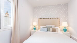 Un dormitorio ordenado con cama, dos mesitas de noche con lámparas, una ventana con cortinas y una pared con estampado.