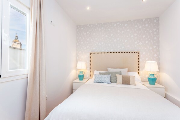 A neatly arranged bedroom with a bed, two bedside tables with lamps, a window with curtains, and a patterned wall.