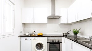 A compact kitchen with a dishwasher, oven, and a small countertop with a kettle and fruits.