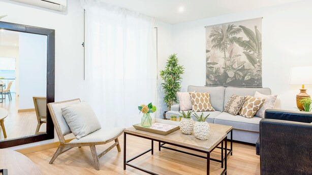 A modern living room with a sofa, coffee table, and chairs. There is a large window with sheer curtains, a potted plant, and a wall-mounted air conditioner.