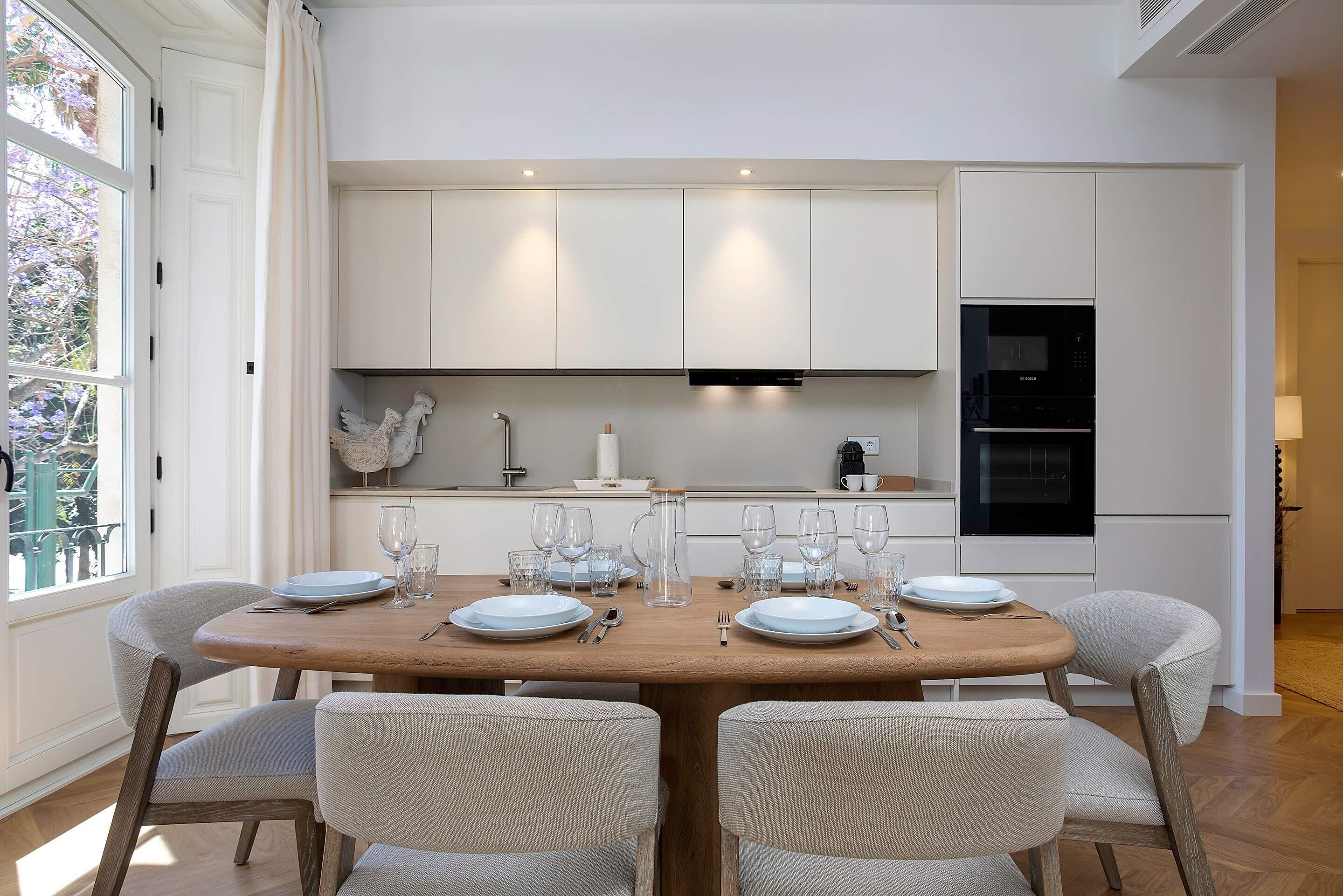 A modern kitchen with white cabinets, a wooden dining table set for four, and a view of the outdoors.