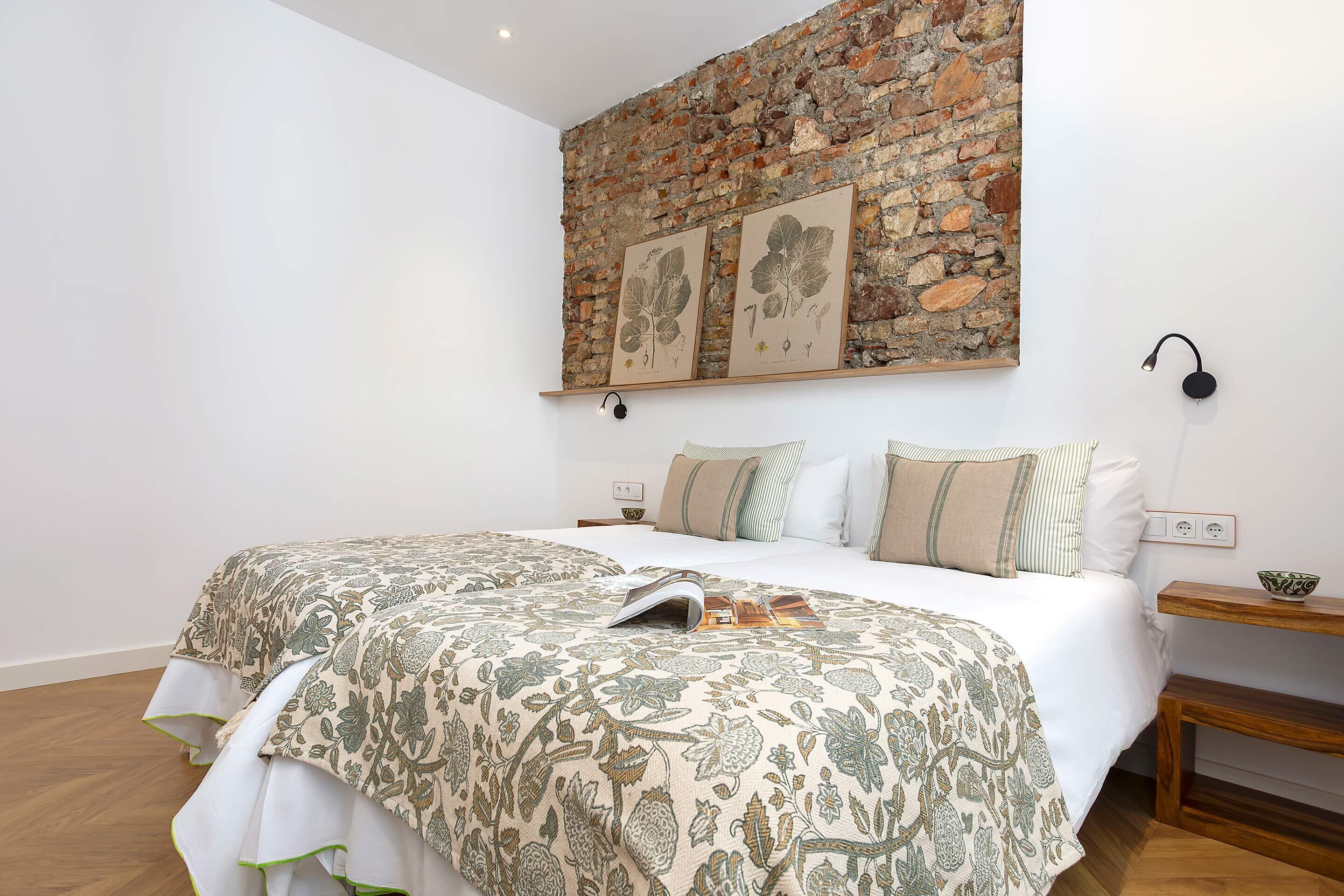 A bedroom with a brick wall, a bed with floral bedding, a nightstand, and a wall-mounted lamp.