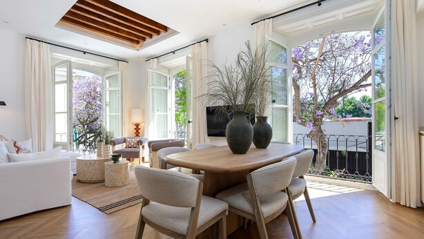 A spacious living area with a round wooden dining table, comfortable seating, and large windows offering views of a garden. - Genteel Home Plaza de la Marina (Málaga)