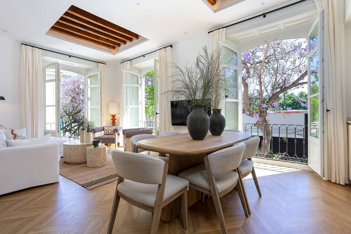 A spacious living area with a round wooden dining table, comfortable seating, and large windows offering views of a garden.