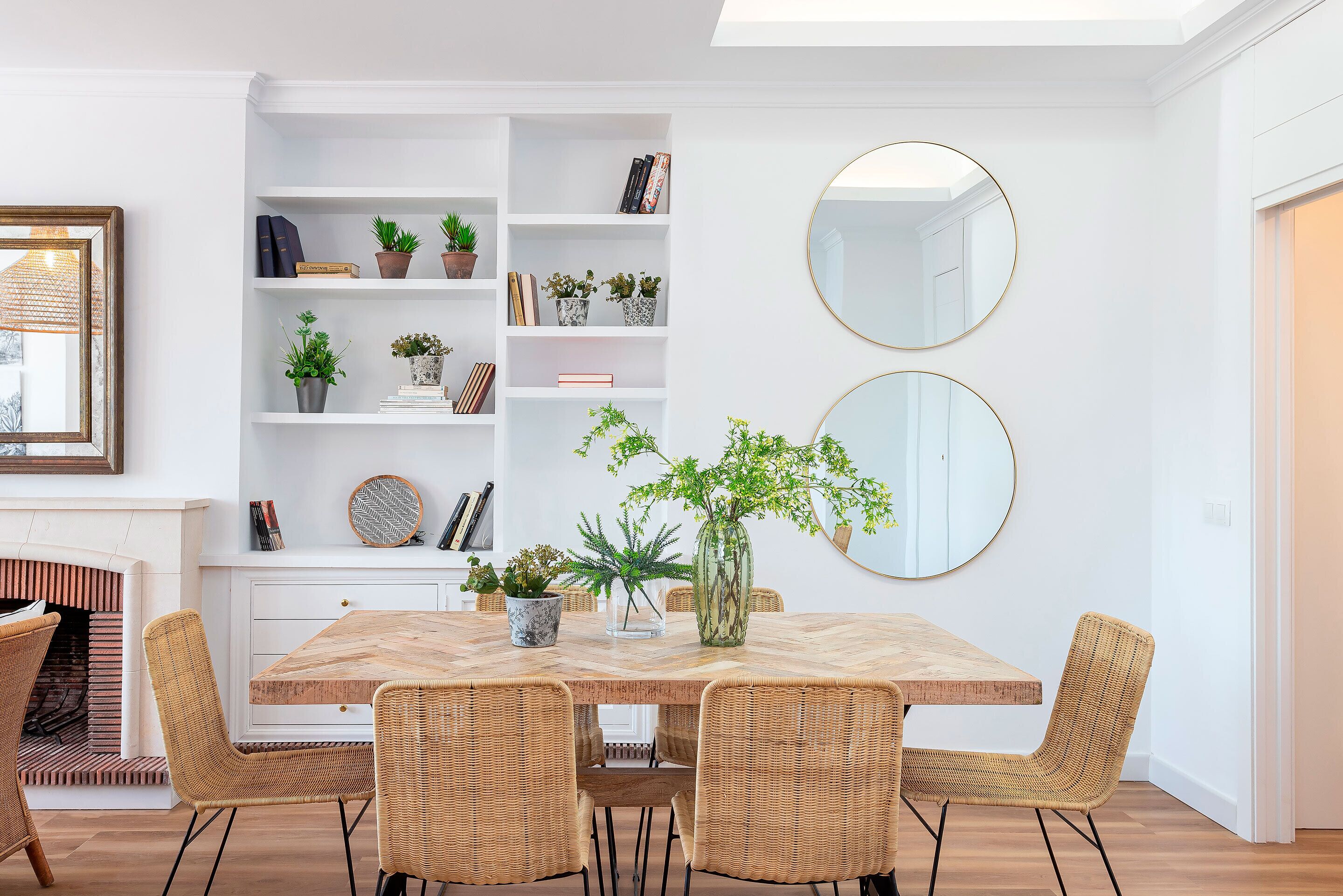 A dining area with a wooden table, wicker chairs, a white bookshelf, and two round mirrors on the wall.