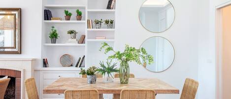 A dining area with a wooden table, wicker chairs, a white bookshelf, and two round mirrors on the wall.