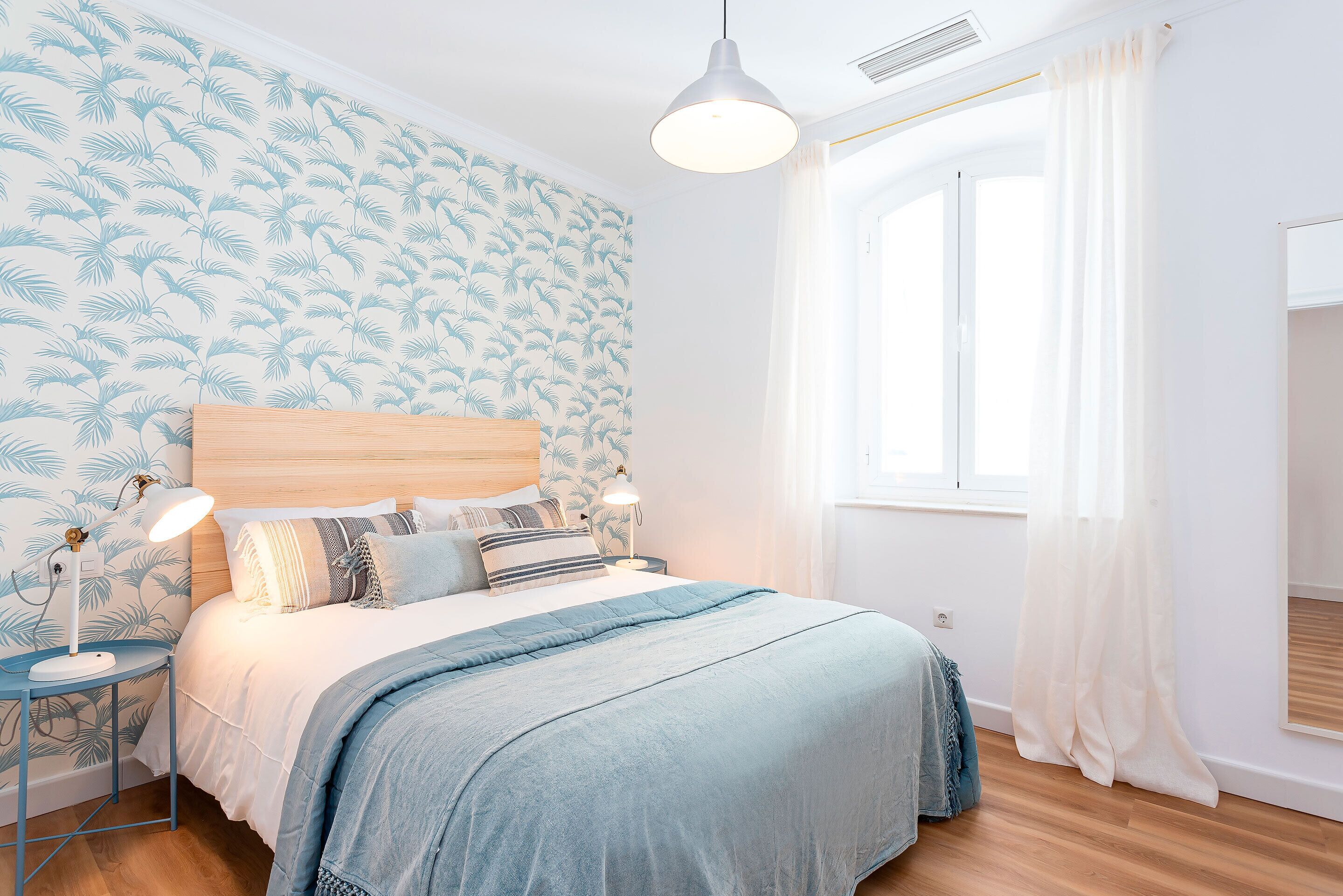 Un dormitorio con cama, mesitas de noche, lámpara, ventana con cortinas y pared con diseño de hojas.
