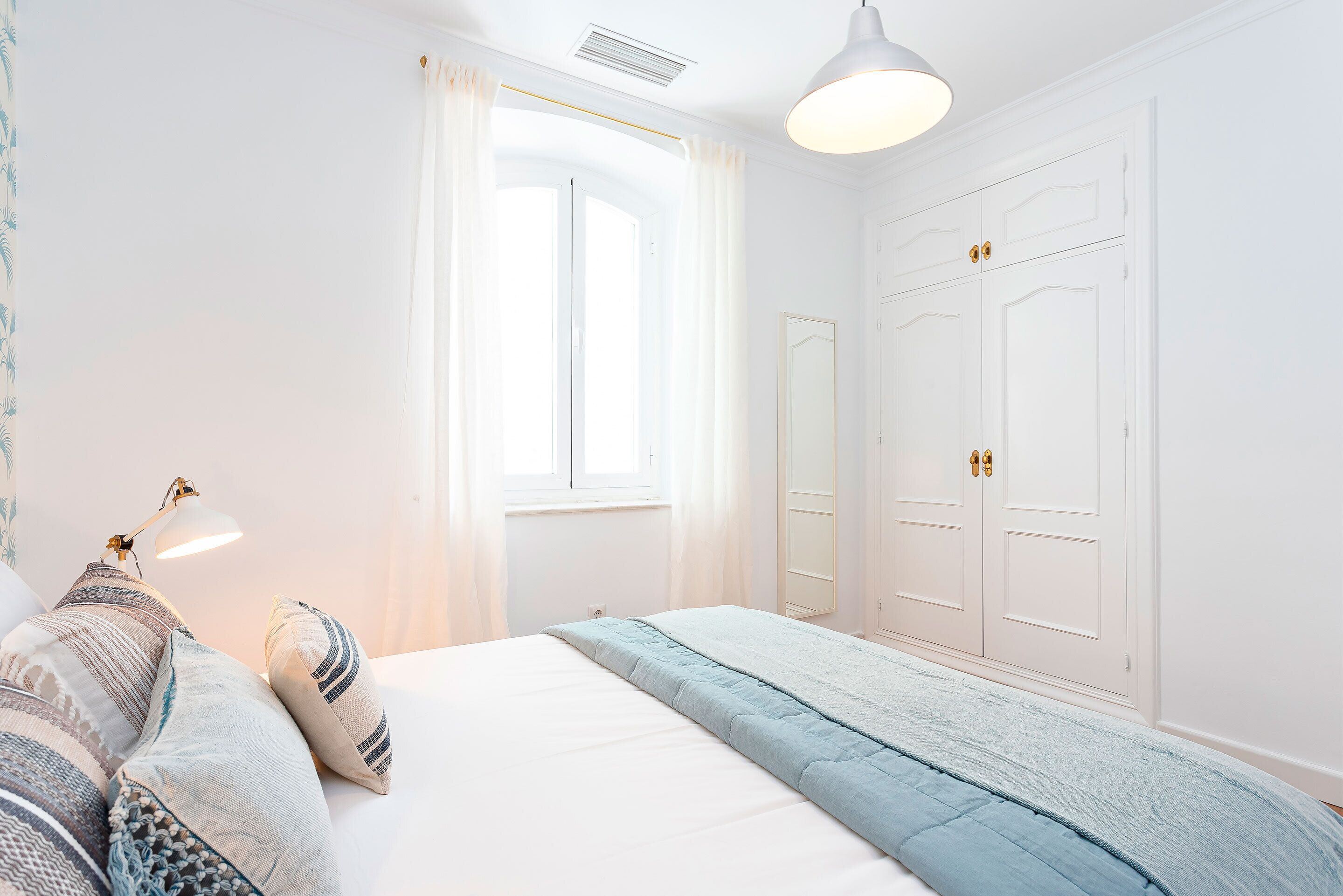 Un dormitorio con cama, lámpara de noche, ventana con cortinas y armario.