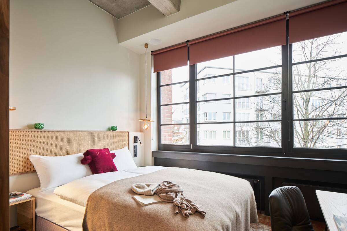 A hotel room with a bed, a window with a view of buildings, a small table, and a chair.