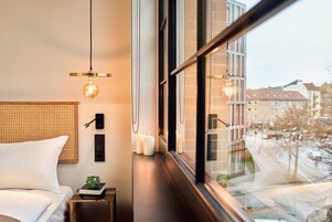 Ein Hotelzimmer mit Bett, Nachttisch, Lampe, Kerzen und Blick auf die Stadt durch große Fenster.