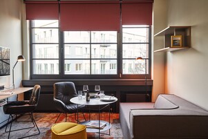 Ein modernes Zimmer mit großem Fenster, einem runden Tisch mit Gläsern und einer Flasche, einer Couch und einem Schreibtisch mit Lampe.