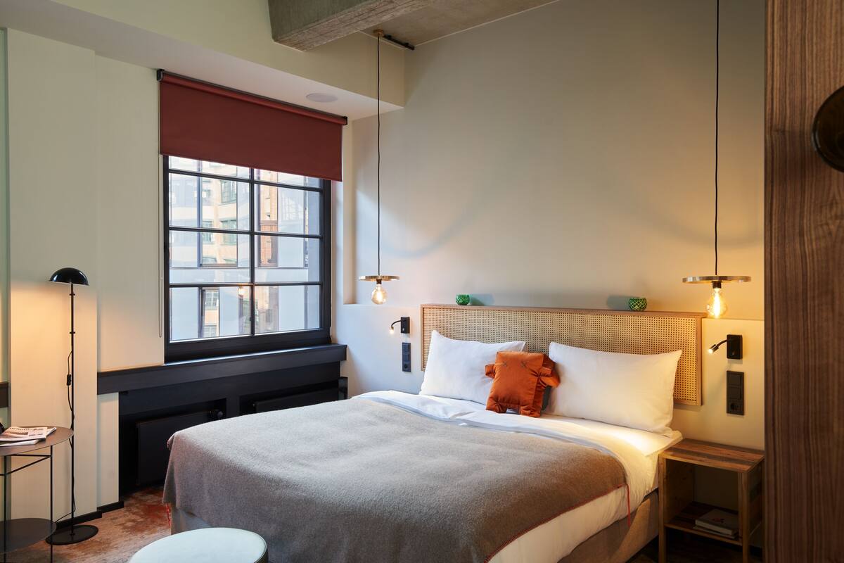 A neatly arranged bedroom with a large bed, a bedside table, a floor lamp, and a window with a view of buildings.