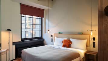 A neatly arranged bedroom with a large bed, a bedside table, a floor lamp, and a window with a view of buildings.
