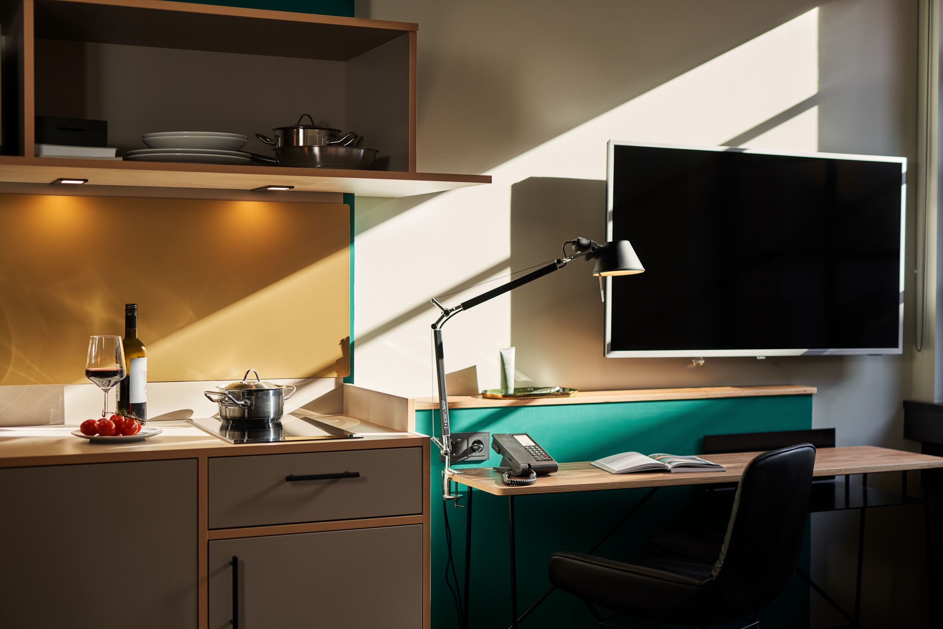 A modern kitchen with a built-in stove, a sink, and a countertop with a wine glass, tomatoes, and a bottle. There is a desk with a chair, a lamp, and a flat-screen TV mounted on the wall.