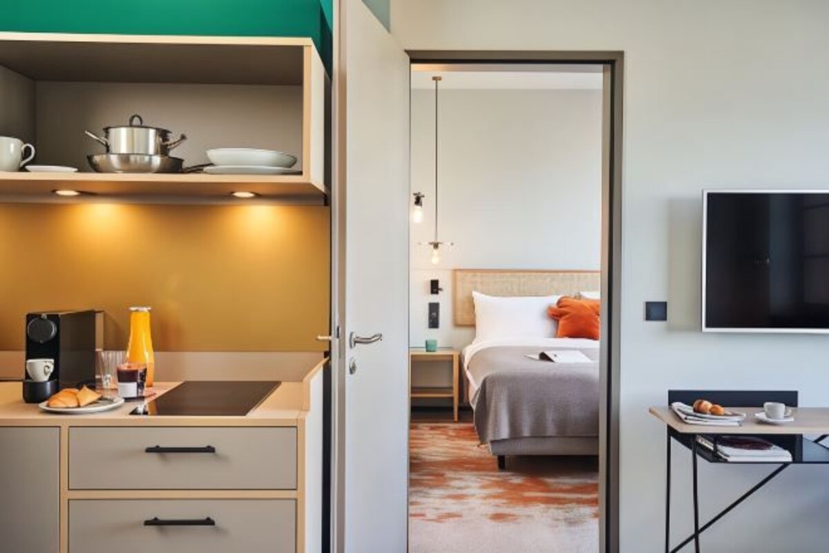 Habitación de hotel con kitchenette, una cama con almohadas, un televisor y una pequeña mesa con una taza y un plato.