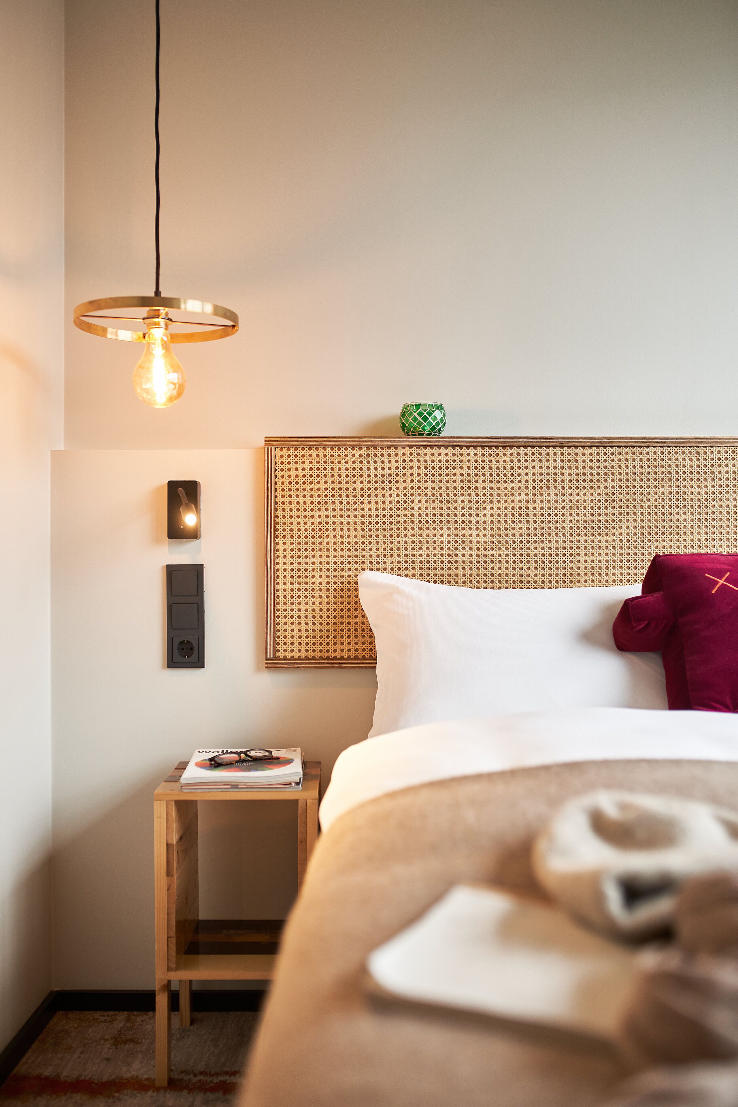Une chambre d’hôtel avec un lit, une table de chevet sur laquelle repose un magazine, un luminaire fixé au mur et une tête de lit en rotin.