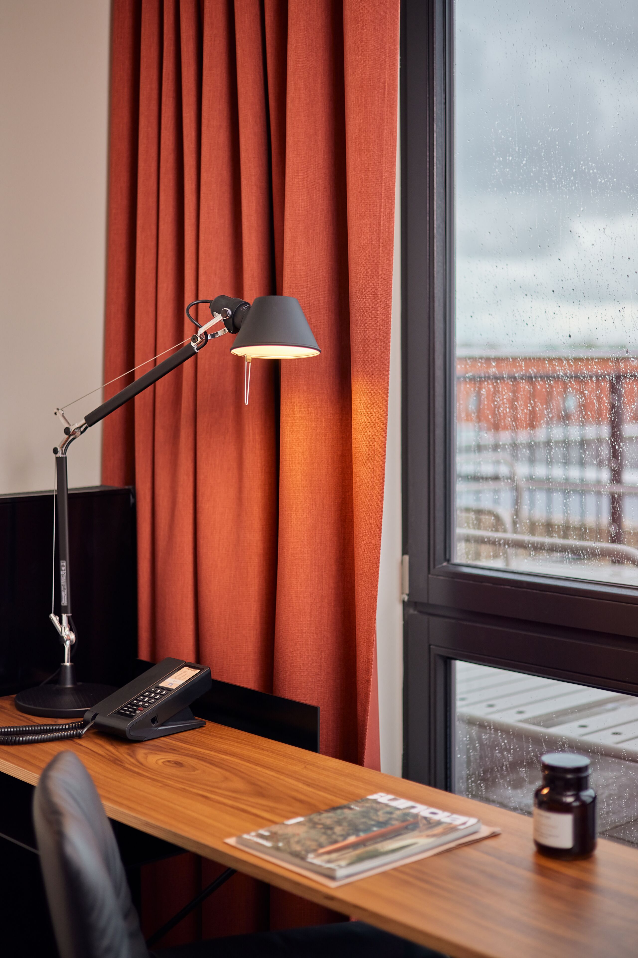 Habitación de hotel con escritorio, lámpara de escritorio, teléfono, una botella y ventana con vista a un edificio.