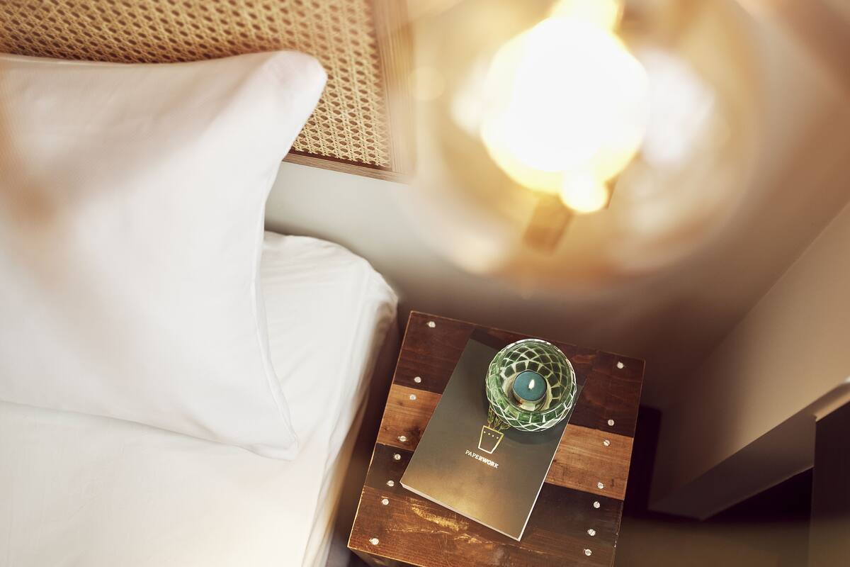 A neatly made bed with a white pillow, a wooden bedside table with a book titled "JAPAN WORK," and a green glass candle holder with a lit candle.