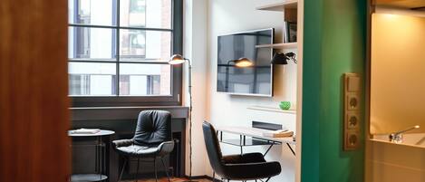 A modern, compact room with a large window, a desk, two chairs, a standing lamp, and a flat-screen TV mounted on the wall.