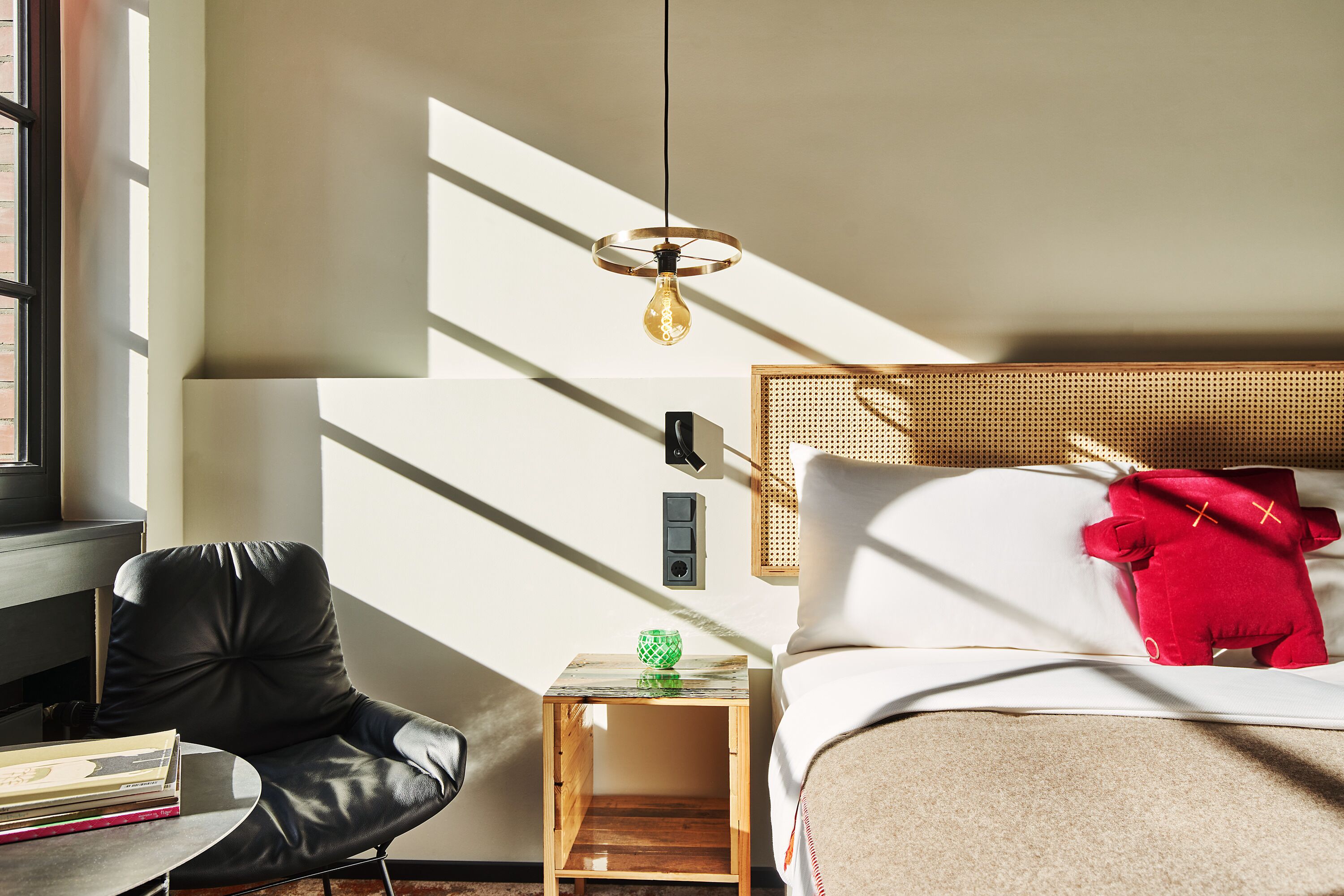 A modern hotel room with a bed, a small wooden nightstand, a black leather chair, and a hanging light fixture.