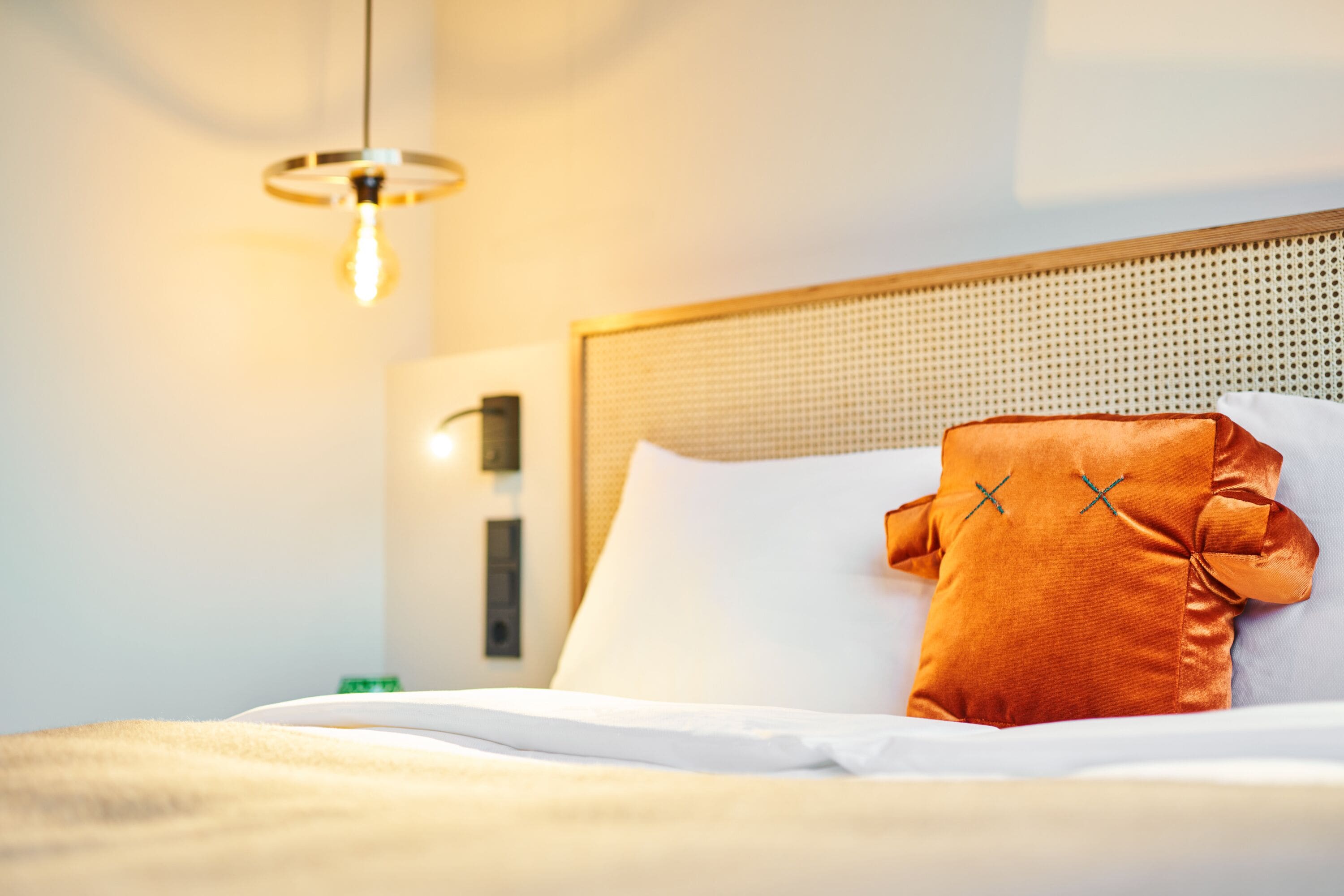 A neatly made bed with white and orange pillows, a woven headboard, and a hanging light fixture.