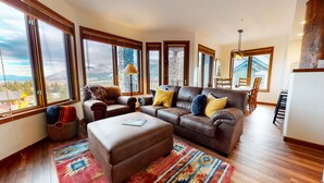 Un salon spacieux avec un canapé en cuir, un pouf et des tapis aux couleurs vives. De grandes fenêtres offrent une vue sur les montagnes.