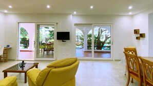 Ein geräumiges Wohnzimmer mit einem gelben Sofa, einem Couchtisch mit Glasplatte und einem Holzesstisch mit Stühlen. An der Wand ist ein Fernseher angebracht und ein großes Schiebeglasfenster führt in einen Außenbereich mit einer Hängematte und einem Esstisch.