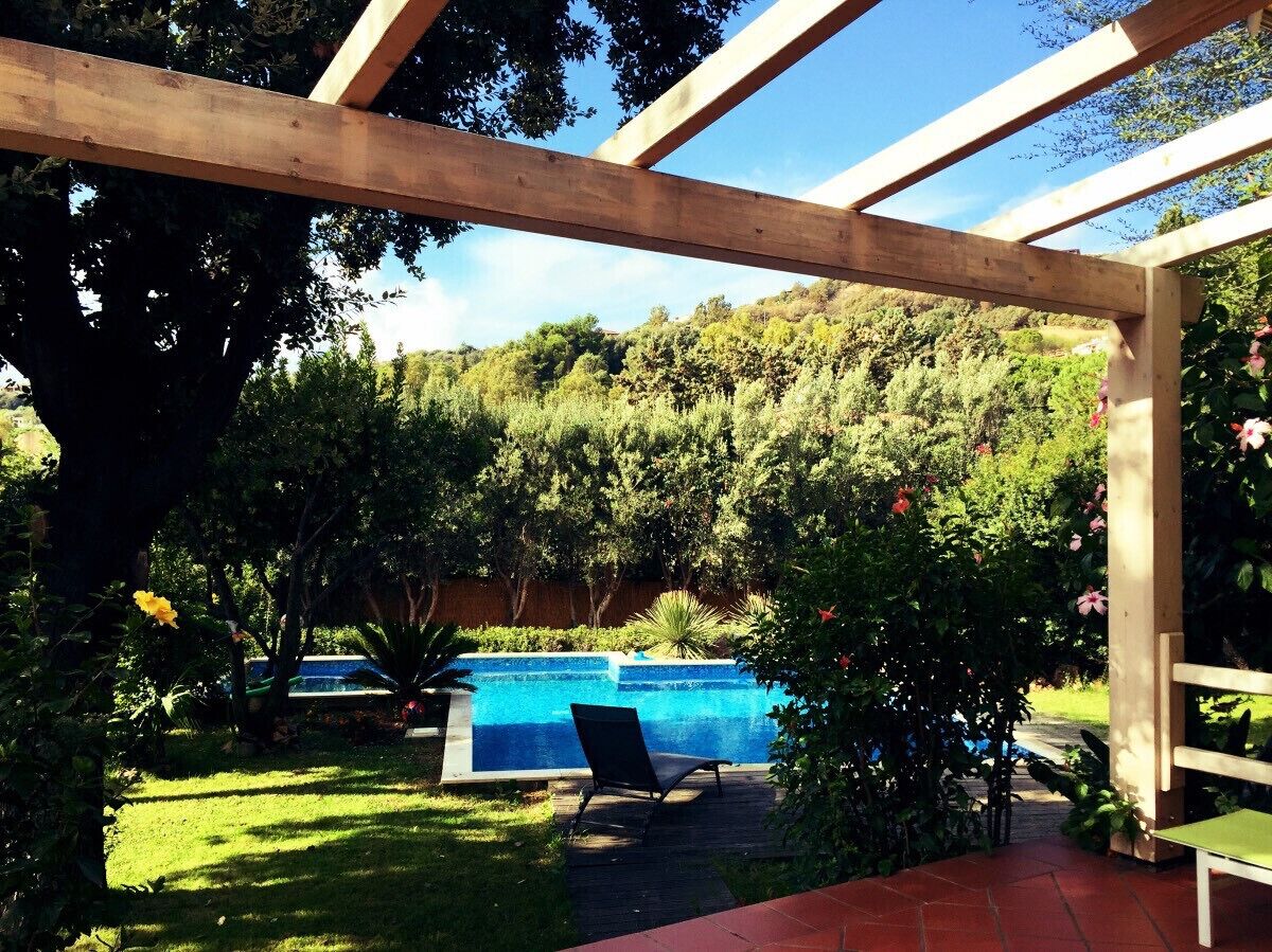 A wooden pergola structure with a pool and a chair in a garden setting.
