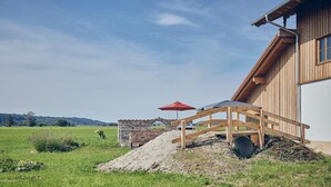 Ein Holzhaus mit einer Steinstruktur davor, einer hölzernen Bank und einem roten Sonnenschirm.