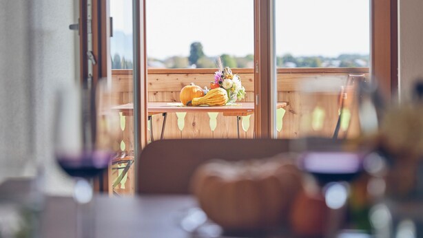 Ein Essbereich mit Holztisch und Stühlen, Blick durch große Fenster und eine Tischdeko mit Kürbis und Blumen.