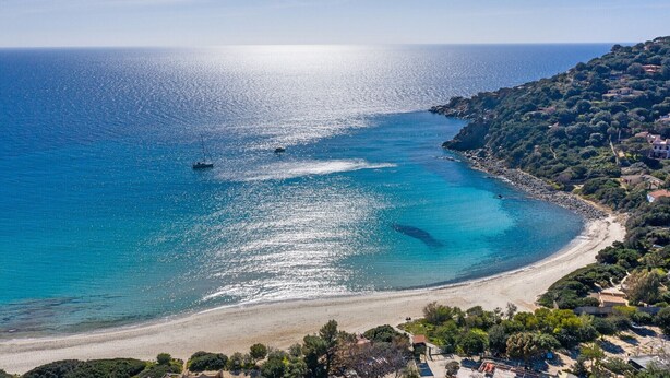 Un paisaje costero con una playa, aguas azules cristalinas y una zona montañosa con vegetación y edificios.