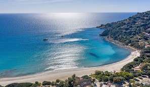 A coastal landscape with a beach, clear blue waters, and a hilly area with vegetation and buildings.