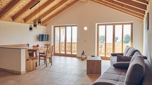 Een ruime woonkamer met een hoge plafond, houten balken, een eettafel met stoelen, een bank en grote ramen.