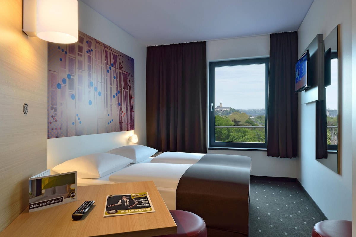Hotelkamer met een bed, een bureau met een afstandsbediening en een tijdschrift, een aan de muur bevestigde televisie en een raam met uitzicht op groen en een gebouw in de verte.