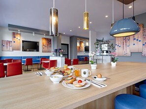 Un espace repas moderne avec une longue table en bois, des chaises rouges et une variété d’aliments pour le petit-déjeuner.