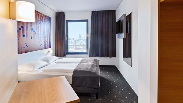 Een hotelkamer met een bed, een kleintje tafeltje, een bankje, een televisie en een raam met gordijnen.