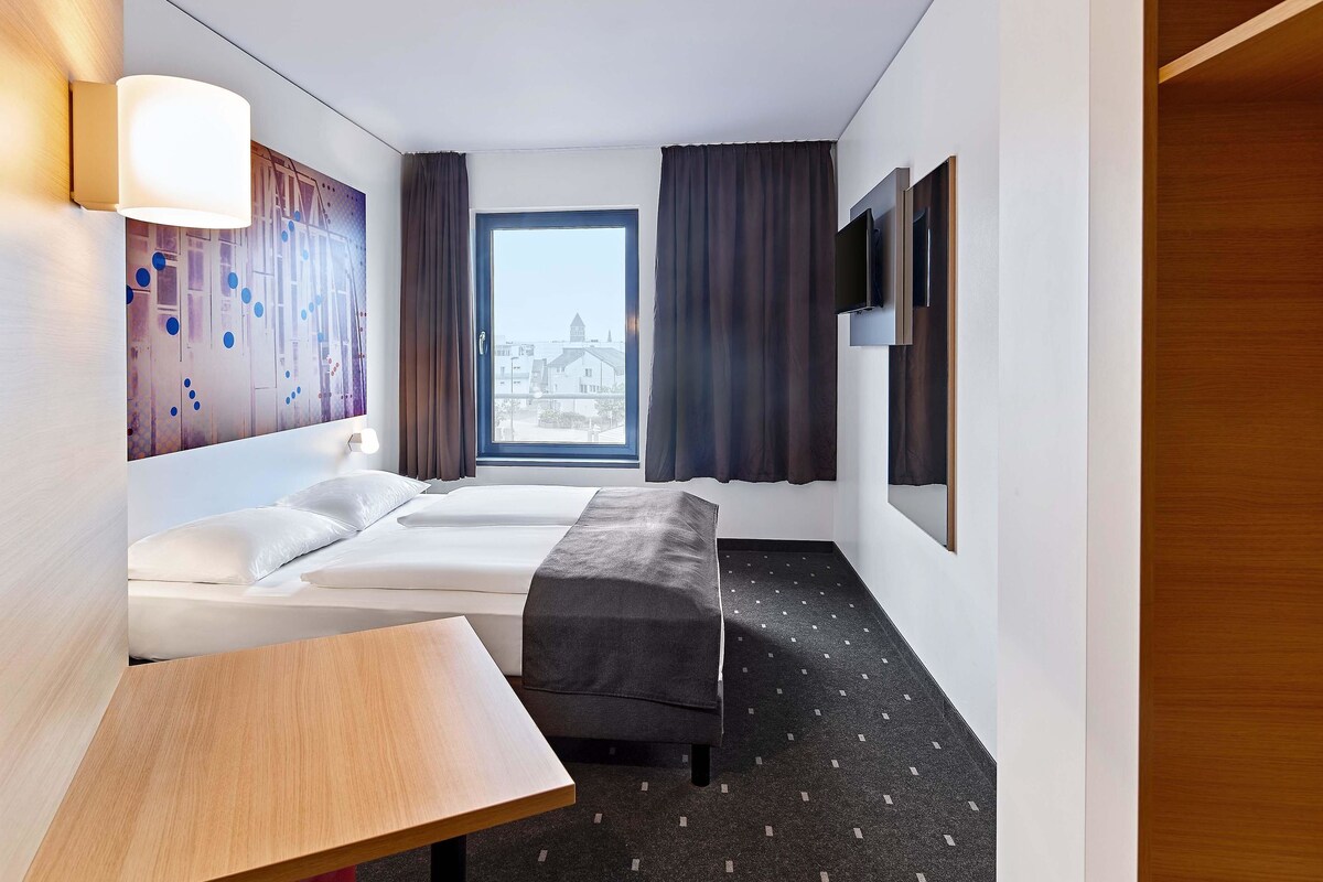 Une chambre d’hôtel avec un lit, une petite table, un banc, une télévision et une fenêtre avec des rideaux.