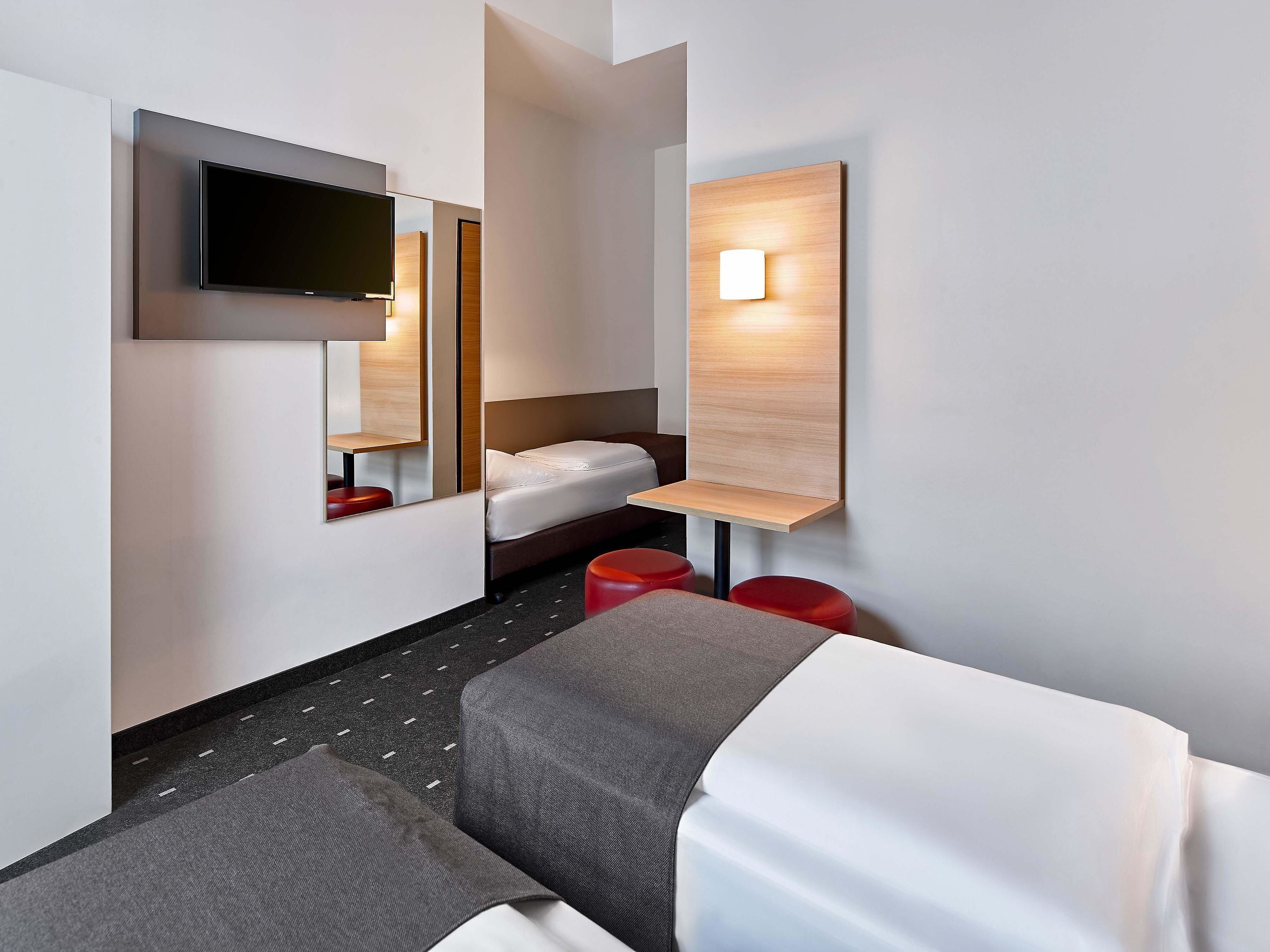 Ein modernes Hotelzimmer mit einem Bett, einem an der Wand montierten Fernseher, einem Nachttisch und einer kleinen Sitzecke mit einem roten Hocker.