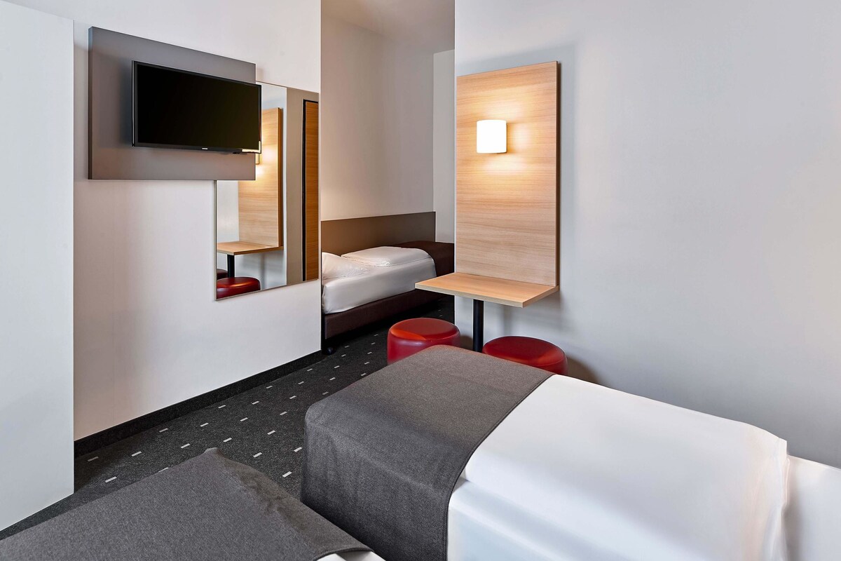 Une chambre d’hôtel moderne équipée d’un lit, d’un téléviseur fixé au mur, d’une table de chevet et d’un petit coin salon avec un tabouret rouge.