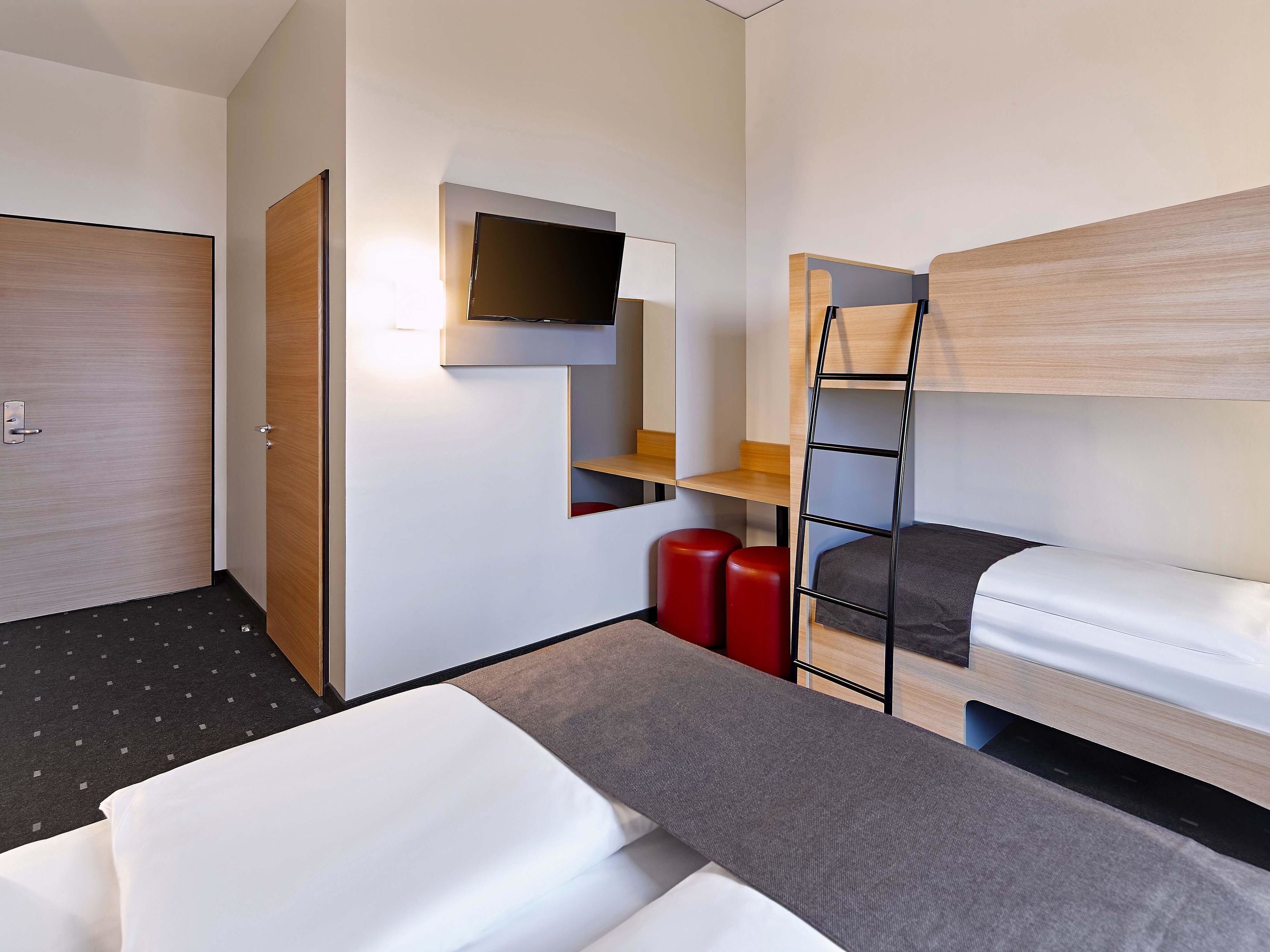 A modern hotel room with a bunk bed, a desk with a chair, a flat-screen TV mounted on the wall, and a small seating area with stools.