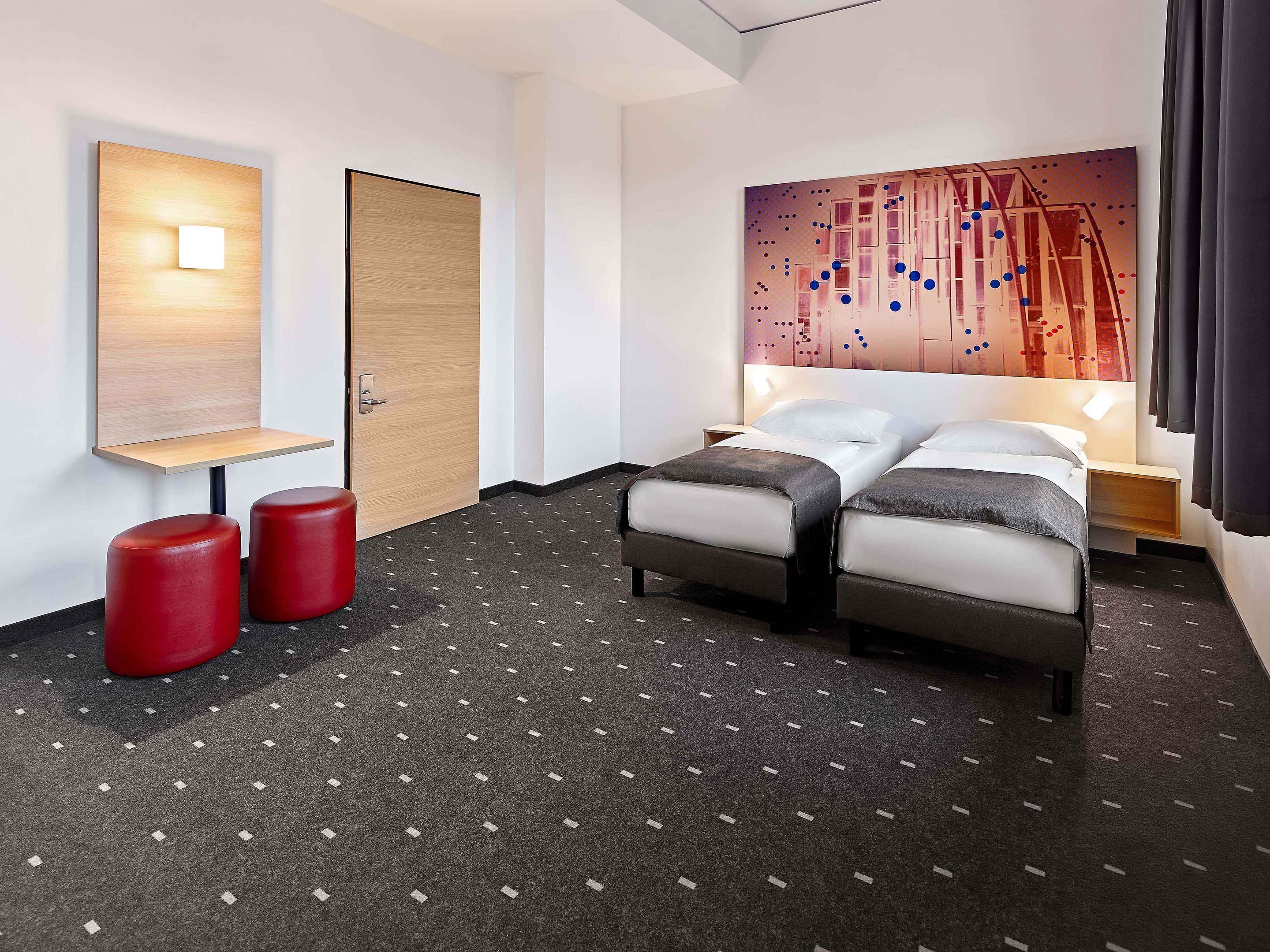 Ein Hotelzimmer mit einem Bett, zwei roten Hocker, einer Holztür, einer Wandleuchte und einem Gemälde.