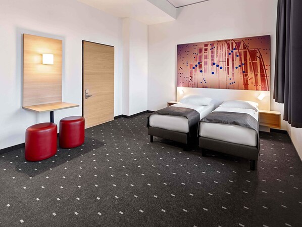 Habitación de hotel con cama, dos taburetes rojos, una puerta de madera, una lámpara de pared y un cuadro.